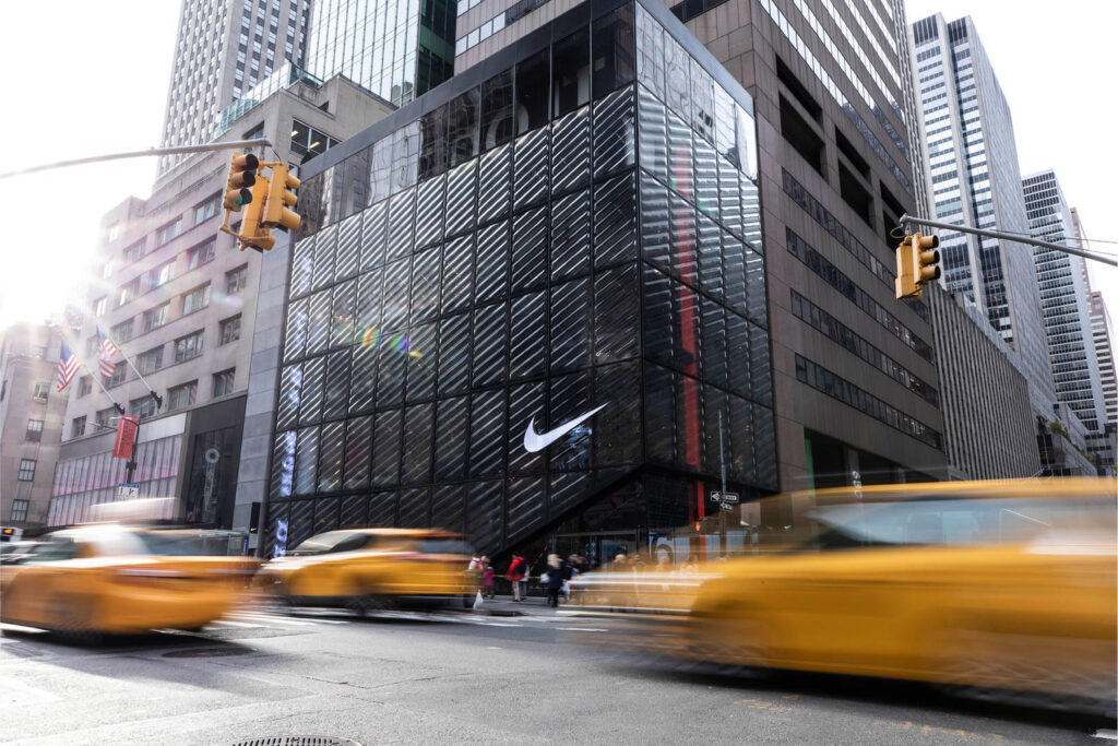 Exterior of Nike House of Innovation in New York City, featuring a striking glass façade with the Nike swoosh logo, surrounded by urban skyscrapers and yellow taxis.
