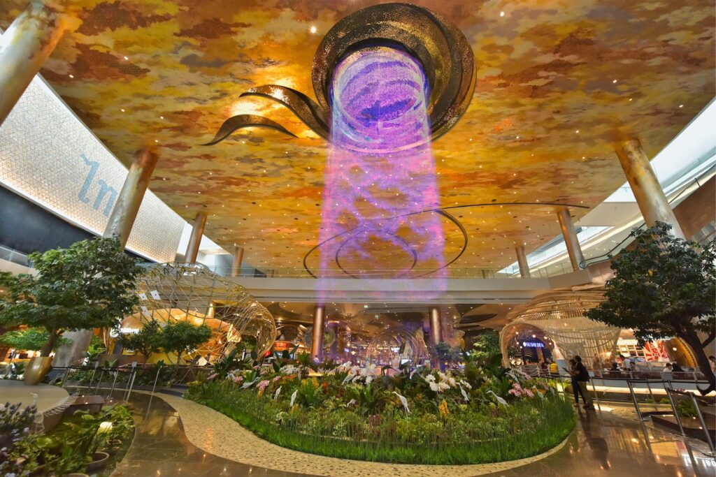 Interior of ICONSIAM’s Alangkarn Waterfall Atrium, featuring a digital waterfall installation cascading from a sculptural ceiling, surrounded by lush greenery and organic architectural forms.