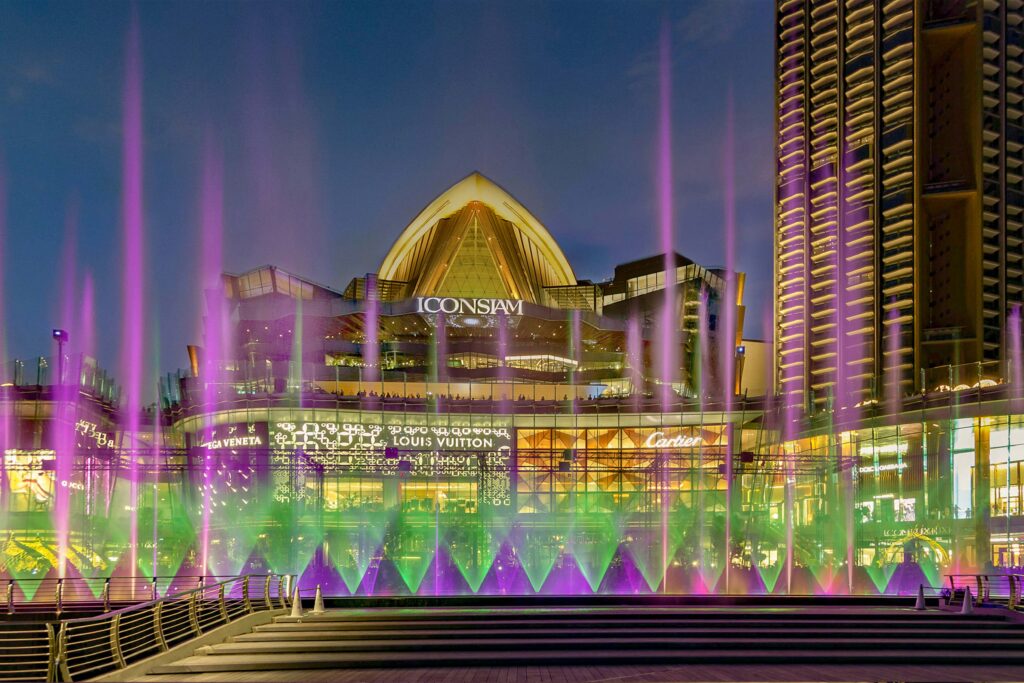 Night view of ICONSIAM’s riverfront façade illuminated with purple and green dancing fountains, highlighting luxury brand storefronts such as Louis Vuitton and Cartier.