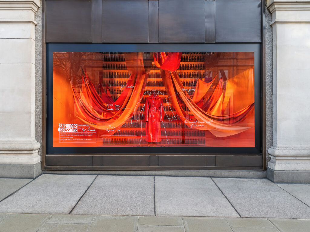 Vibrant red Selfridges Obsessions window display inspired by hot sauce, with draped fabrics and fashion styled in bold fiery tones.