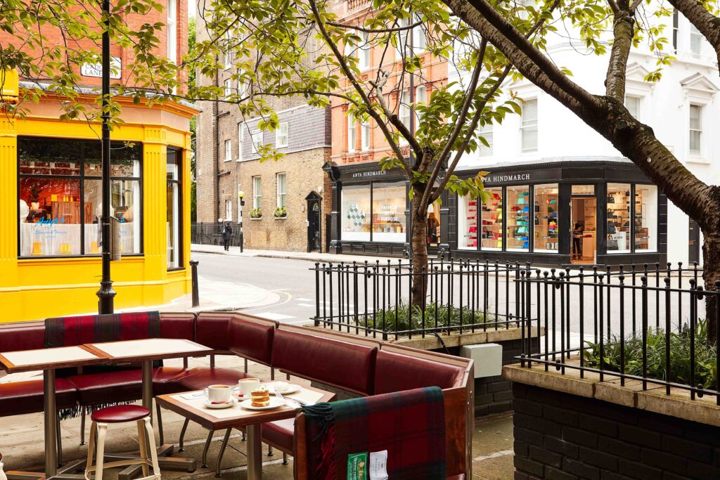 Street view of Anya Café in London’s Chelsea, with outdoor seating under a tree and a view of the bright yellow café façade beside the black storefronts of Anya Hindmarch on Pont Street.