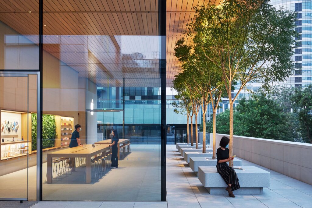 Interior and exterior view of Apple Sanlitun in Beijing, featuring minimalist glass architecture, an open courtyard with trees, and modern seating areas.
