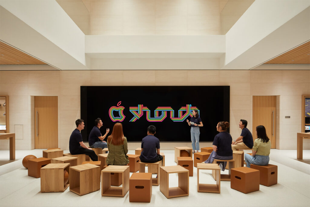 An Apple Store interior featuring a minimalist design with light wood and neutral tones. A group of people is seated on wooden cube stools in front of a large black digital screen displaying a colorful, stylized Apple logo. A presenter stands beside the screen leading a session, creating a collaborative and creative atmosphere.