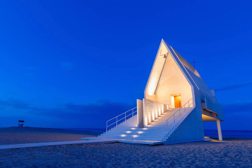 Minimalist beachfront chapel with triangular geometry, illuminated stairway, and a striking presence against the evening blue sky.