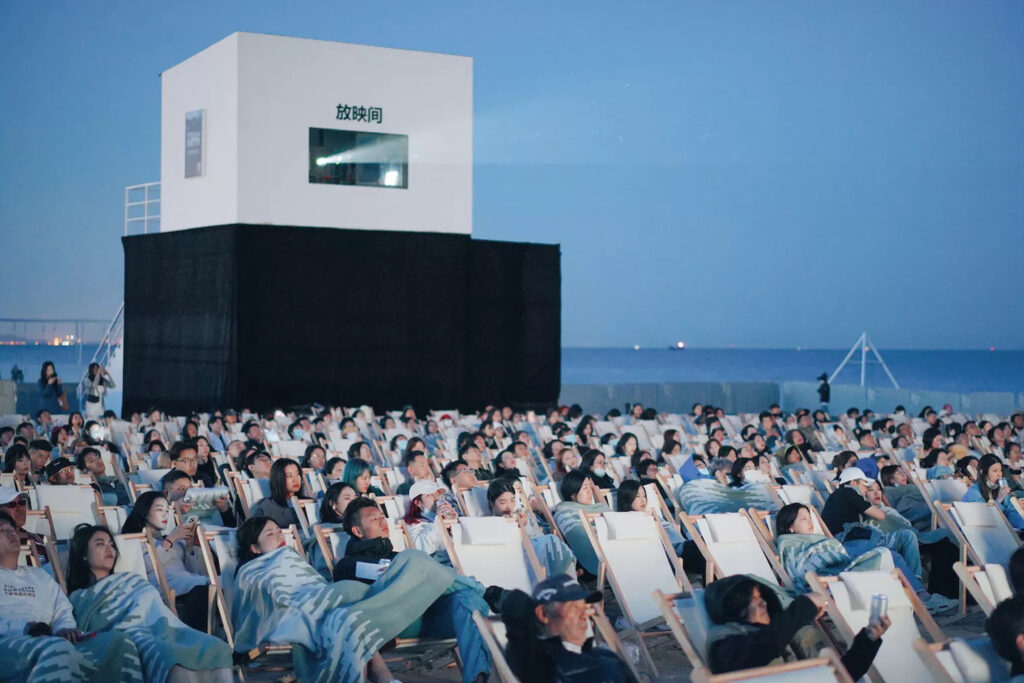 Open-air beach cinema at dusk with audiences seated in deck chairs facing the sea, creating a communal cultural experience.