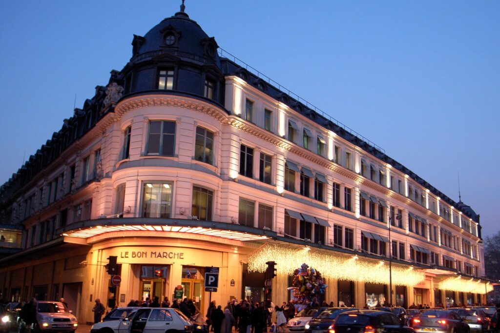 Exterior of Le Bon Marché department store in Paris at dusk, illuminated with warm lights and festive decorations along the historic building façade.