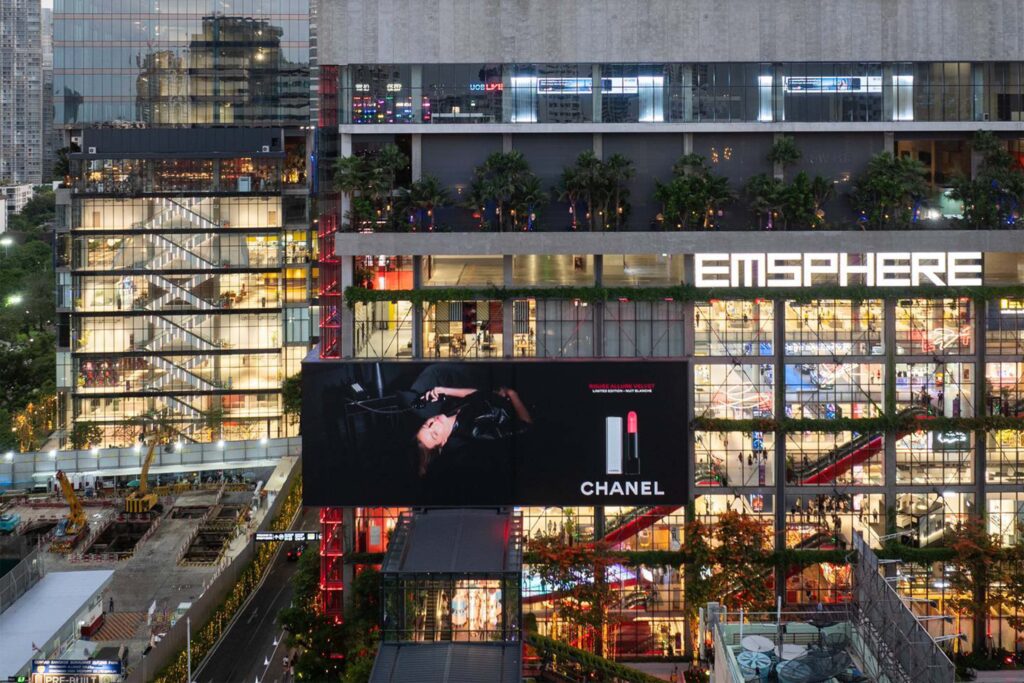 Exterior view of Emsphere Bangkok at dusk, showcasing the modern glass and concrete façade illuminated by retail lighting and signage. A large Chanel billboard dominates the center, while the multi-level structure reveals open terraces, greenery, and visible escalators inside the shopping complex.