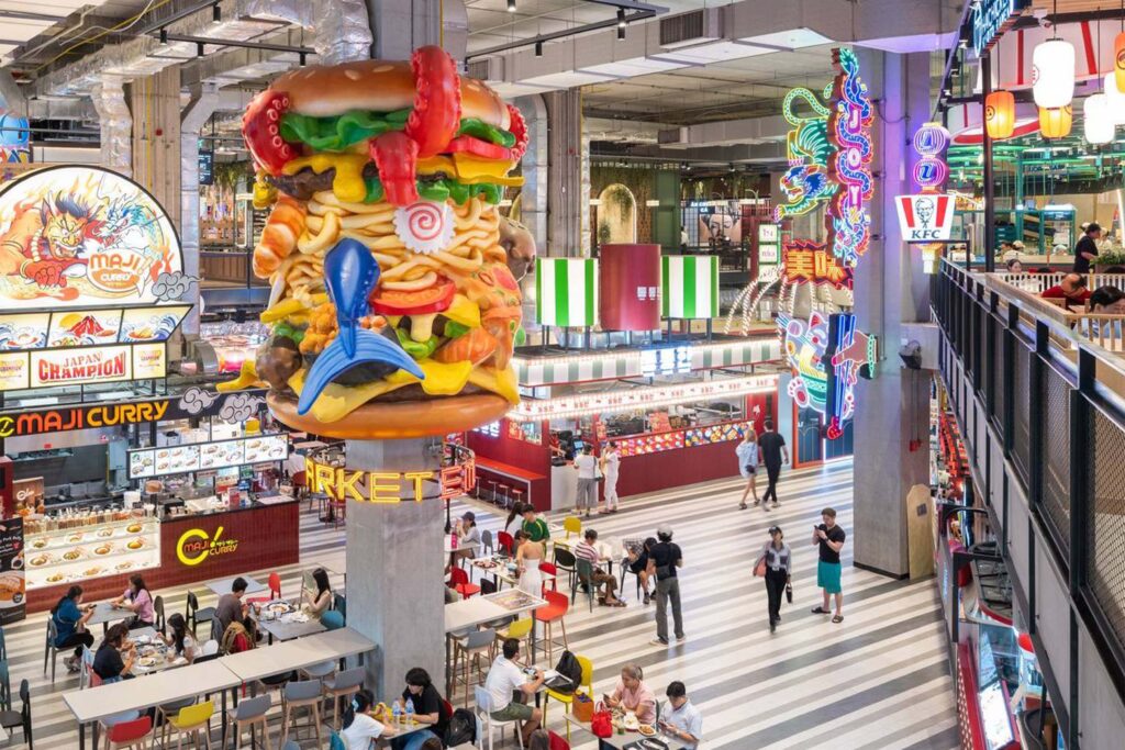 Vibrant food market inside Emsphere Bangkok showcasing playful oversized sculptures, neon signage, and diverse dining options in a dynamic, urban setting.