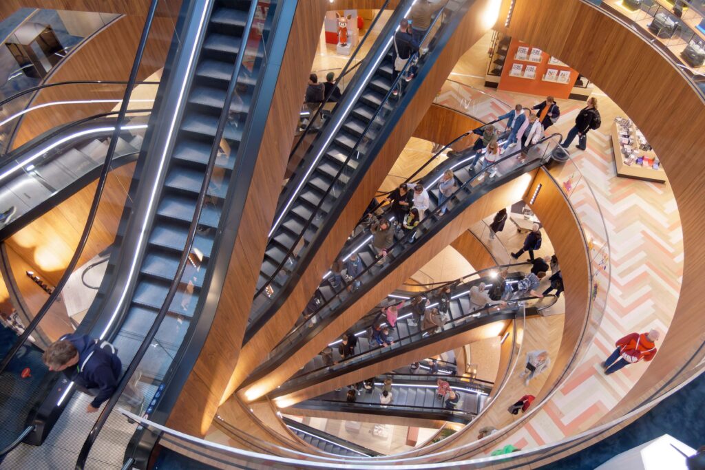Dynamic interior view of KaDeWe department store showing a dramatic spiral of wooden escalators connecting multiple floors, with shoppers moving between modern retail levels under warm architectural lighting.