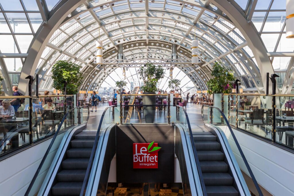 Bright interior of Le Buffet Wintergarten restaurant featuring a glass-domed ceiling, lush indoor plants, and escalators leading up to a spacious dining area filled with natural light and contemporary furnishings.