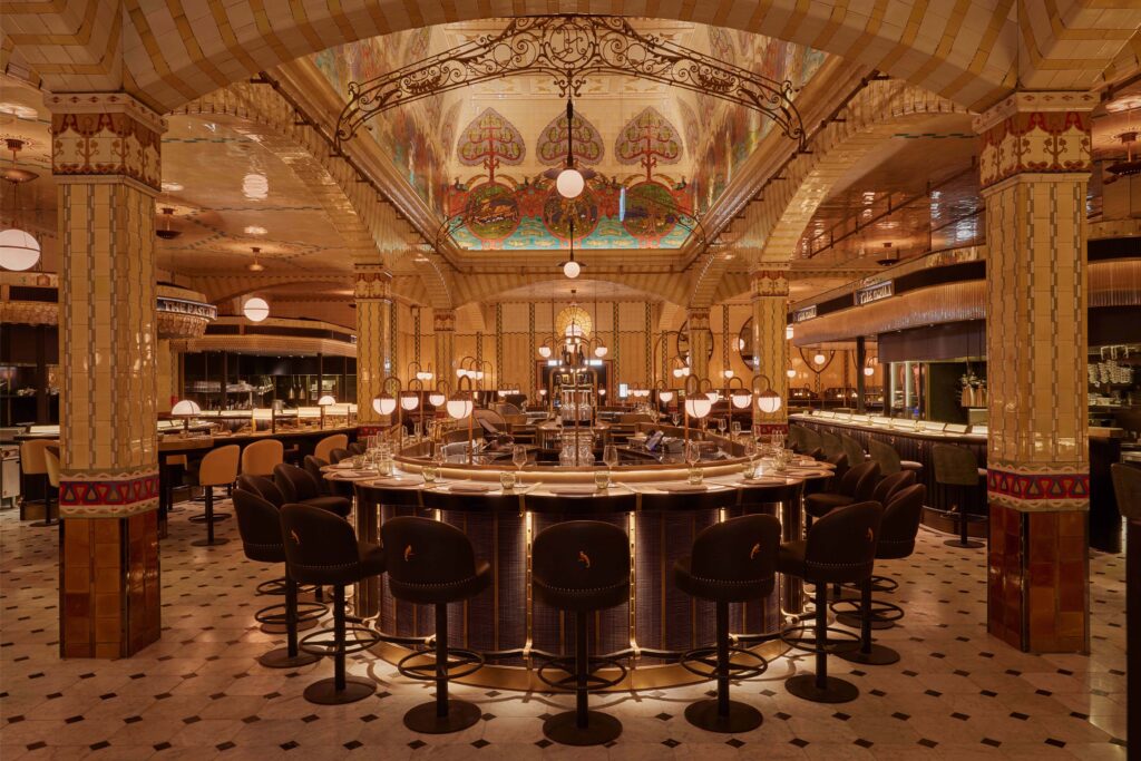 The Art Deco-inspired Harrods Dining Hall in London, showcasing ornate tiled arches, a central circular bar with ambient lighting, and decorative ceiling murals, blending heritage craftsmanship with contemporary dining elegance.