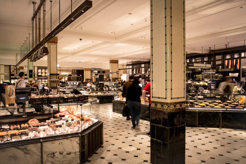 Interior of the Harrods Food Hall in London, featuring elegant tiled columns, marble counters, and delicatessen displays filled with meats, cheeses, and pastries, creating a luxurious market-style atmosphere within the historic department store.