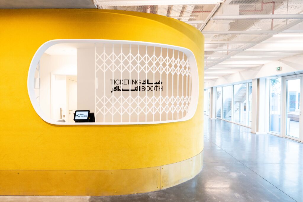 Ticketing booth at Hayy Cinema, featuring a bright yellow curved wall and minimalist Arabic-English signage within Art Jameel’s contemporary design.