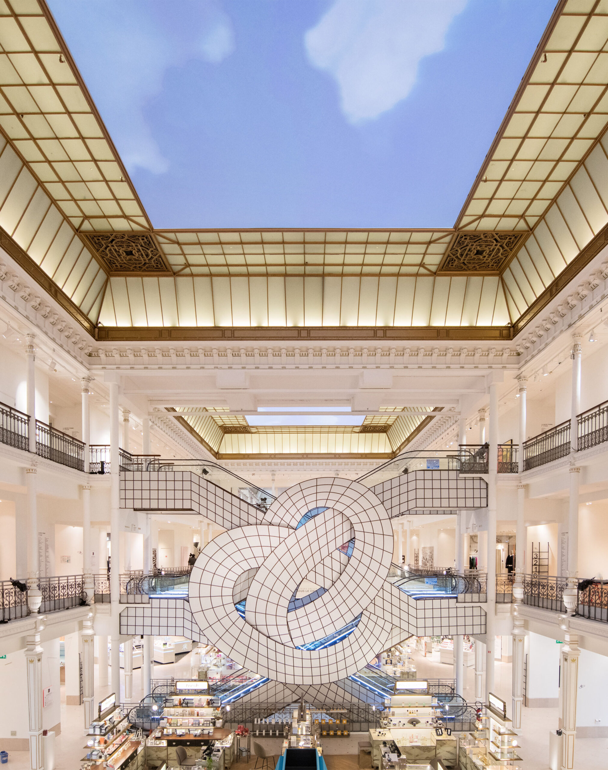 Leandro Erlich’s large-scale installation at Le Bon Marché in Paris, featuring an intricate sculptural knot made of tiled panels suspended within the grand atrium, surrounded by the store’s elegant neoclassical architecture, glass ceilings, and retail displays below.