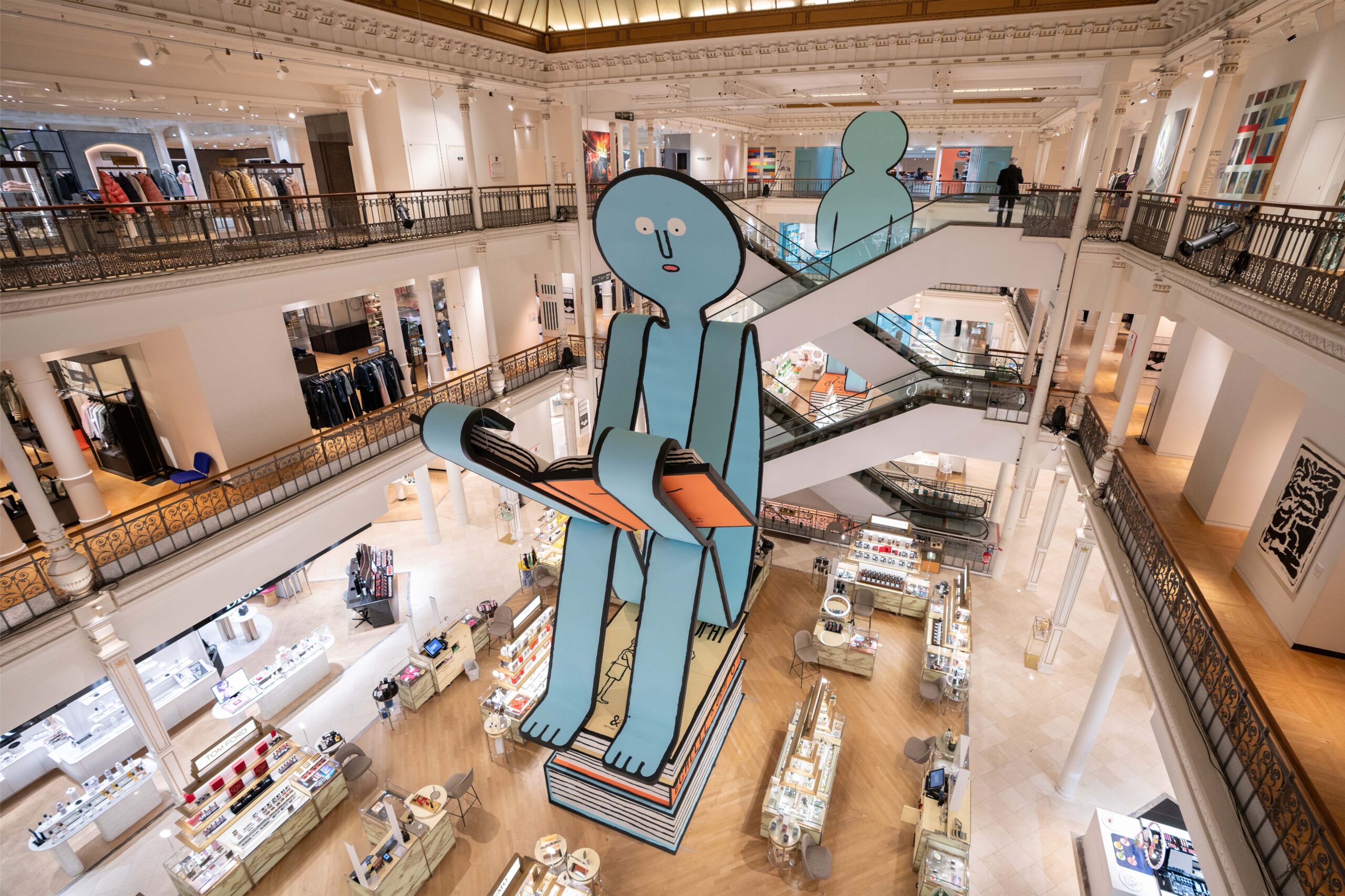Installation inside Le Bon Marché in Paris featuring a large blue figure reading an oversized book, part of a contemporary art display integrated within the department store’s iconic neoclassical interior, grand staircases, and luxury retail environment.