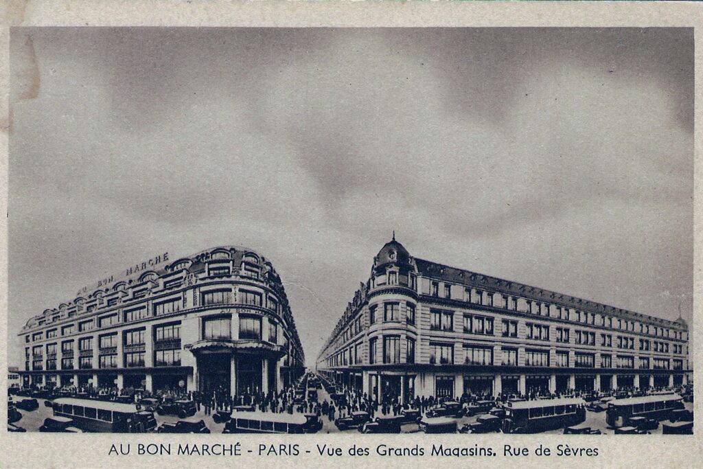 Historic black-and-white photograph of Au Bon Marché department store in Paris, showing the grand façades of the two main buildings on Rue de Sèvres, with early 20th-century cars and crowds in front.