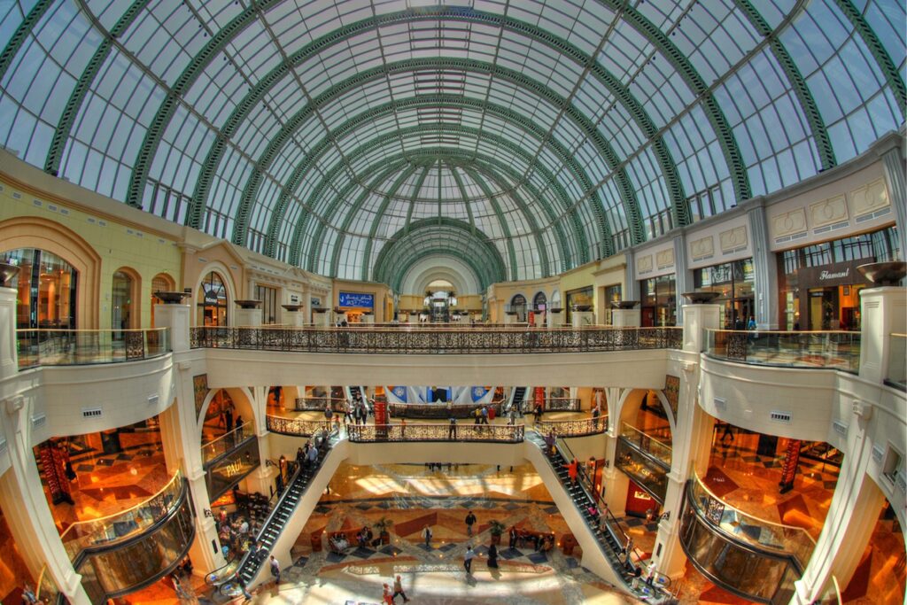 Interior view of the Mall of the Emirates in Dubai featuring a grand glass-domed atrium, multiple retail levels connected by escalators, marble flooring, and luxury brand storefronts illuminated by natural light.
