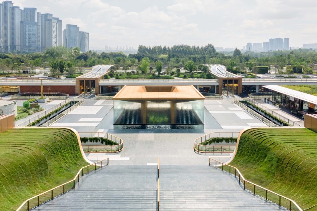 Grand Staircase view of SKP Chengdu showing the central plaza with a sculptural water pavilion surrounded by landscaped mounds and retail pavilions under a bright, open sky.