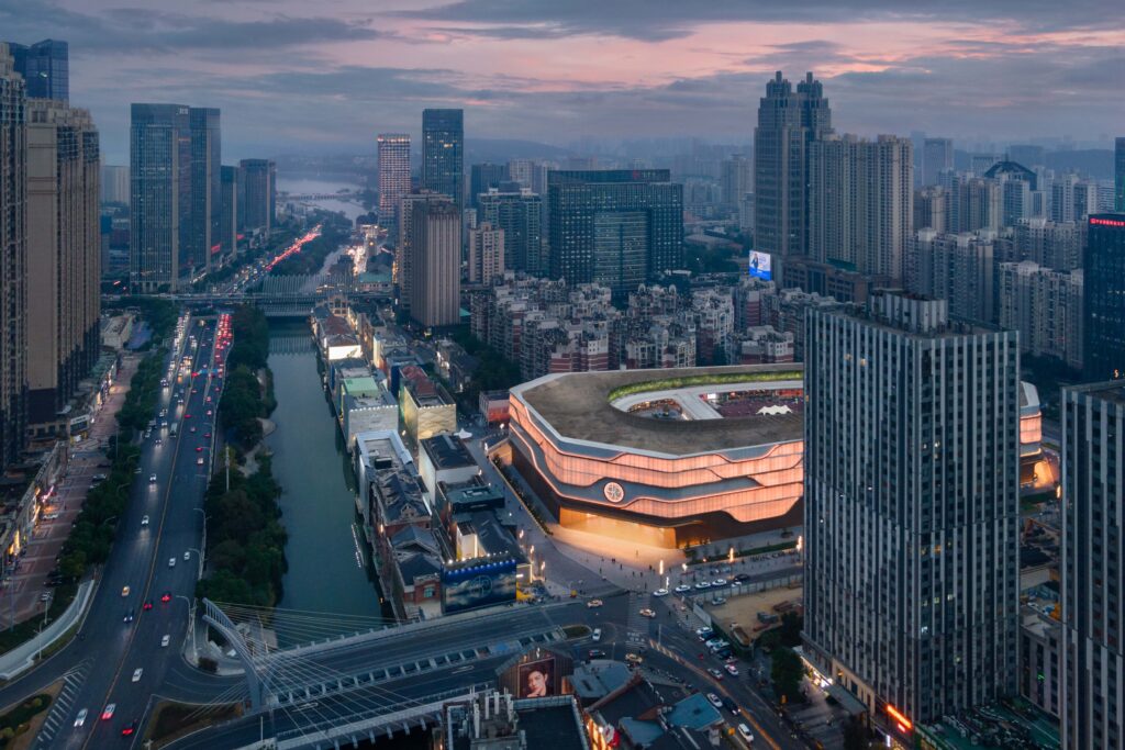 Aerial evening view of SKP Wuhan, highlighting the softly illuminated terracotta-toned façade within the dense urban skyline, adjacent to the canal and main thoroughfare.