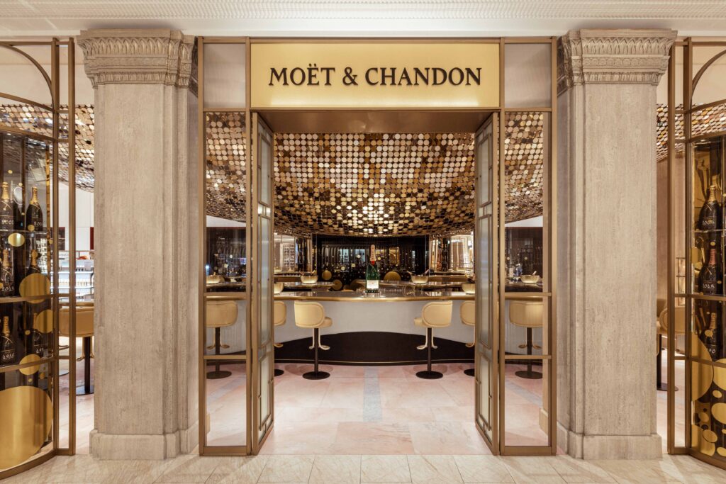 Entrance to the Moët & Chandon Champagne Bar at Harrods, framed by classical columns and golden glass doors, revealing a luminous interior with a sculptural gold ceiling installation and elegant cream seating.