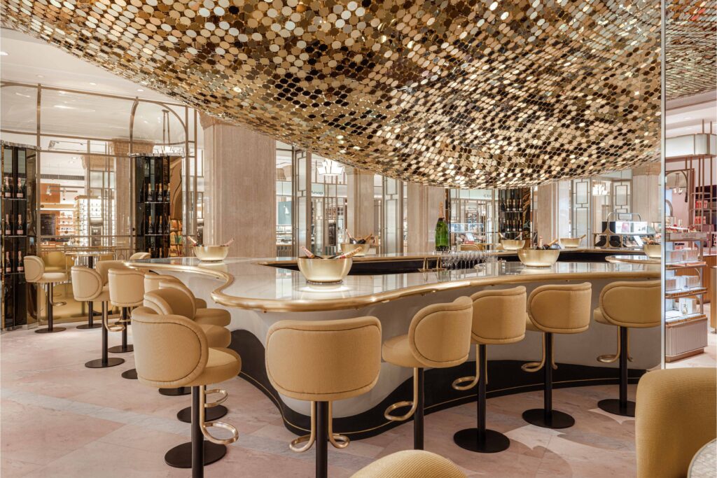 Interior of the Moët & Chandon Champagne Bar at Harrods featuring a curved marble counter surrounded by gold-upholstered bar stools beneath a shimmering canopy of golden metallic discs.