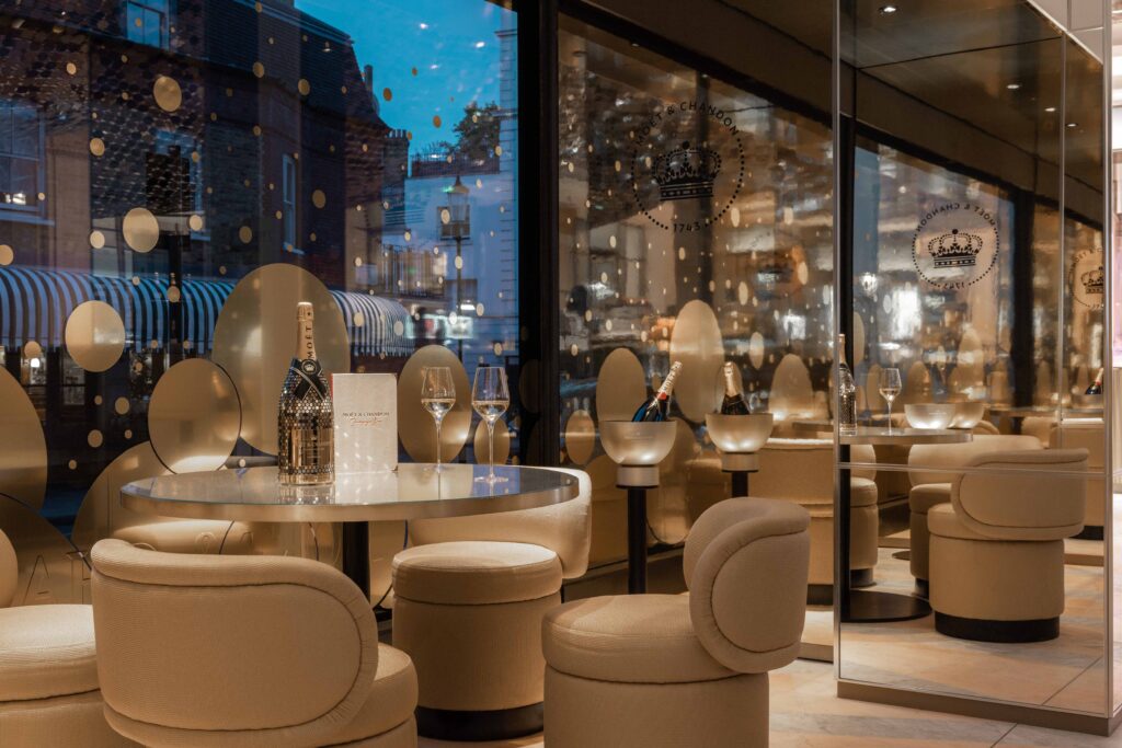 ounge seating area inside the Moët & Chandon Champagne Bar at Harrods with circular tables, champagne displays, and gold bubble motifs on the windows reflecting the London streetscape outside.