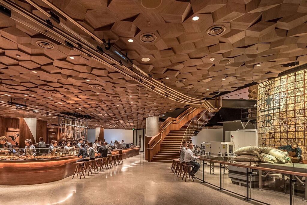 Interior of a contemporary café with a sculptural wooden ceiling, curved coffee bar, and warm ambient lighting. People sit along the bar and staircase in a spacious, modern design setting featuring geometric wood textures and polished concrete floors.