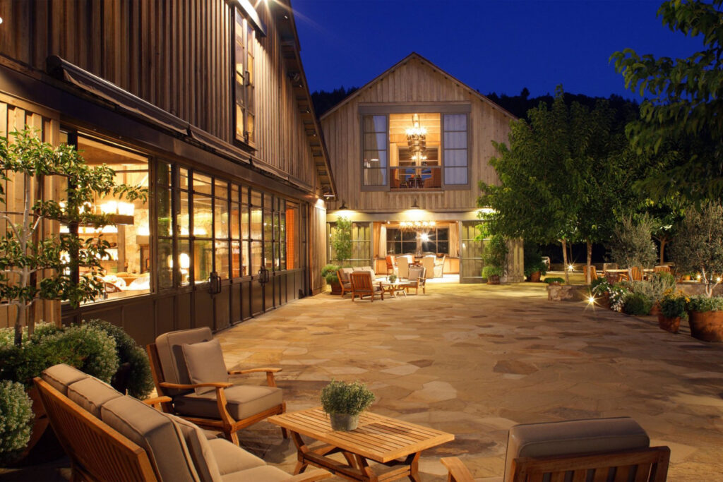 Evening view of The Napa Valley Reserve featuring warm lighting, rustic wooden architecture, and outdoor seating surrounded by trees and stone paving.