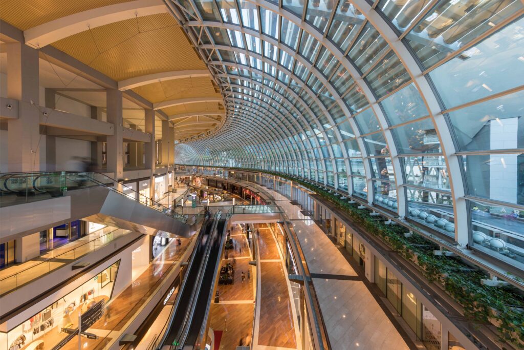 Interior of The Shoppes at Marina Bay Sands in Singapore featuring a curved glass roof, luxury retail stores, multiple escalators, and polished marble floors illuminated by natural light from the expansive skylight structure.