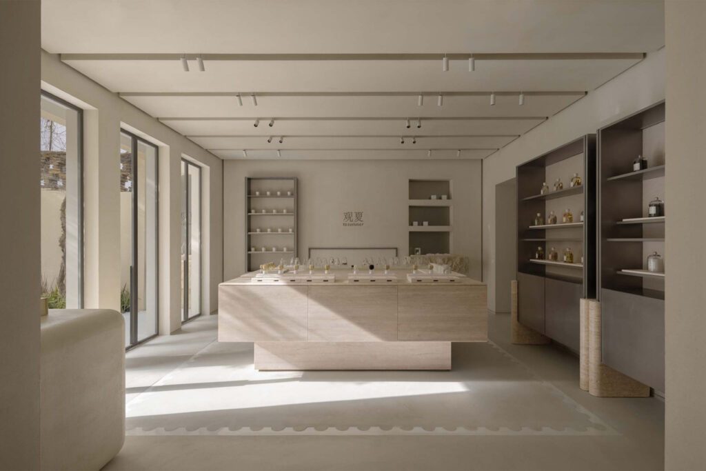 Minimalist interior of the To Summer fragrance boutique featuring a central travertine display table, warm neutral tones, and natural light streaming through full-height windows.