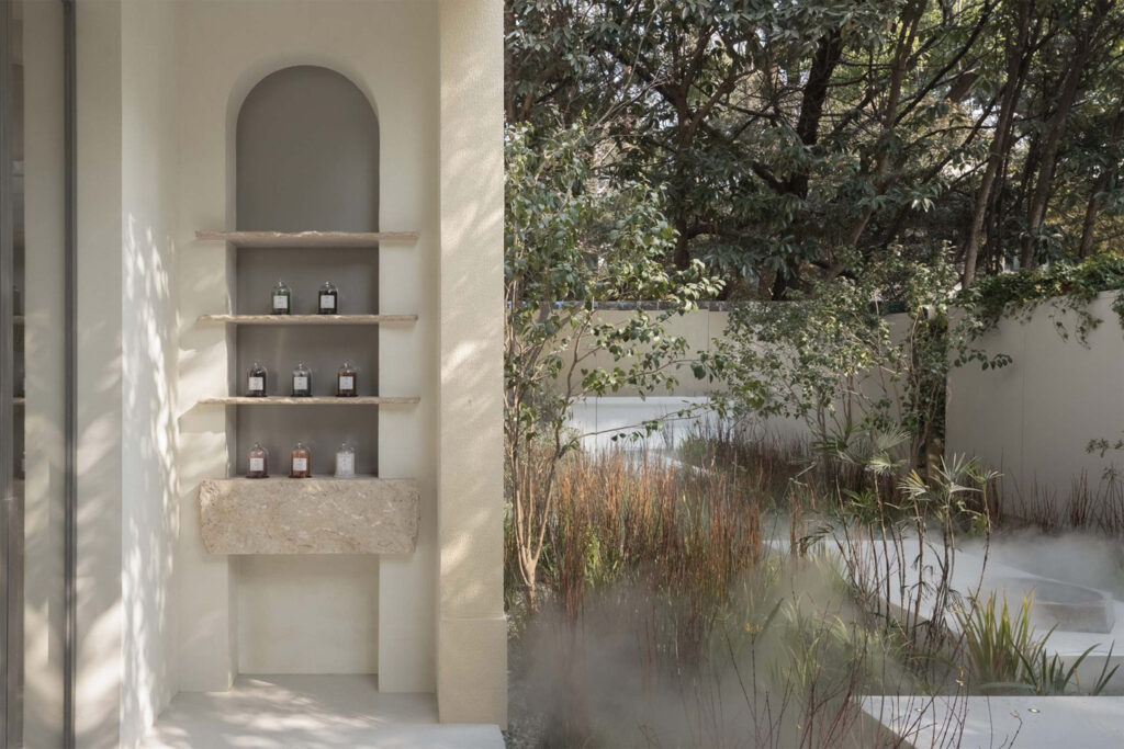 Outdoor display niche at To Summer showcasing glass-domed perfume bottles on stone shelves, surrounded by a tranquil misty garden with natural vegetation.