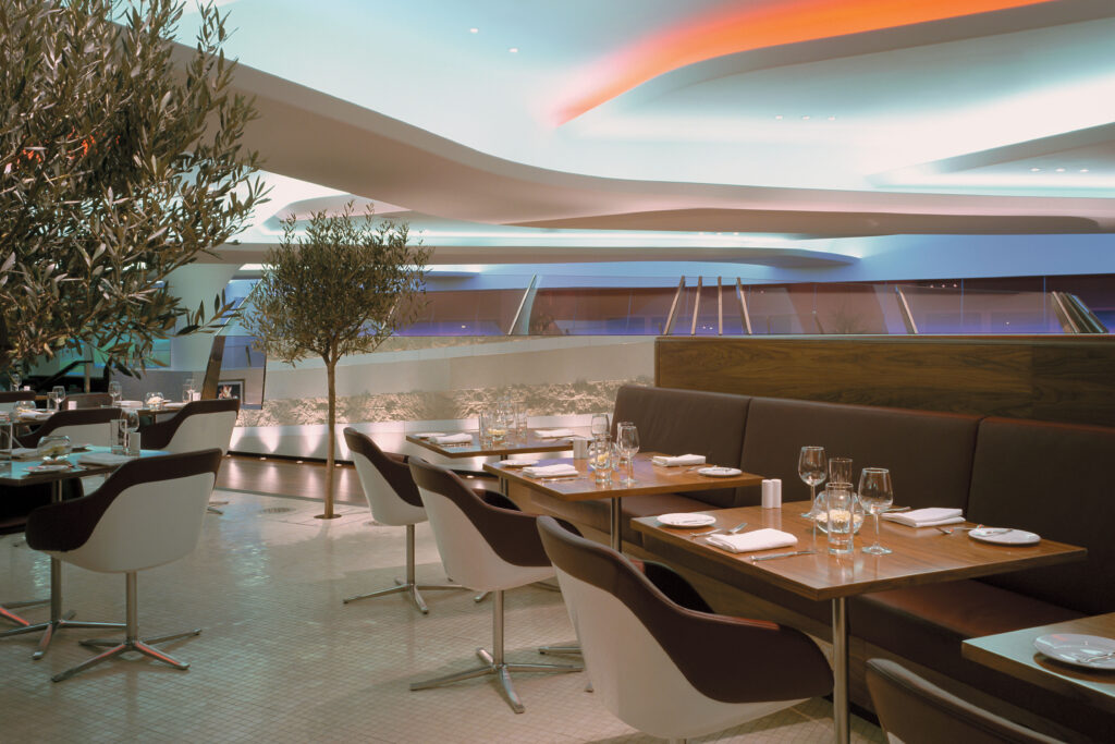 Interior views of the Virgin Atlantic Heathrow Clubhouse in London, designed with a fluid, futuristic aesthetic. The space features organic curves, sculptural ceilings with ambient lighting, and luxurious materials such as wood, leather, and glass. Various zones include a sunken lounge, cocktail bar, restaurant seating with olive trees, and relaxation areas with Eames chairs and large projection screens.