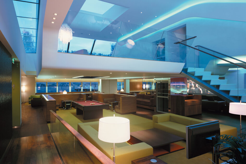 Interior views of the Virgin Atlantic Heathrow Clubhouse in London, designed with a fluid, futuristic aesthetic. The space features organic curves, sculptural ceilings with ambient lighting, and luxurious materials such as wood, leather, and glass. Various zones include a sunken lounge, cocktail bar, restaurant seating with olive trees, and relaxation areas with Eames chairs and large projection screens.