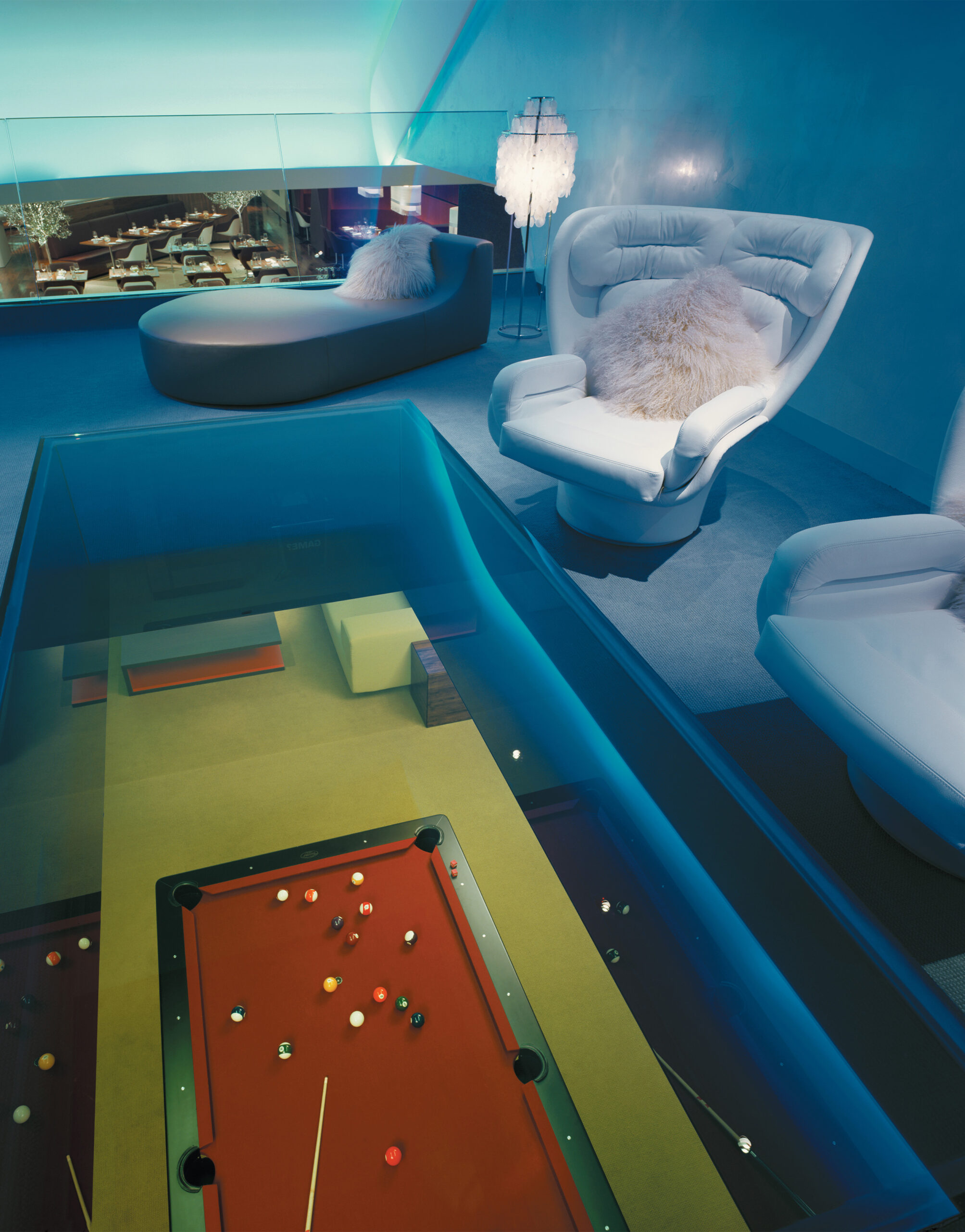 Interior of the Virgin Atlantic Heathrow Clubhouse Skylounge in London, featuring a red pool table viewed through a glass floor, modern white lounge chairs with fluffy cushions, and ambient blue lighting creating a futuristic, relaxed atmosphere.
