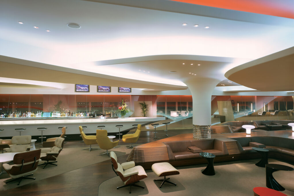 Interior views of the Virgin Atlantic Heathrow Clubhouse in London, designed with a fluid, futuristic aesthetic. The space features organic curves, sculptural ceilings with ambient lighting, and luxurious materials such as wood, leather, and glass. Various zones include a sunken lounge, cocktail bar, restaurant seating with olive trees, and relaxation areas with Eames chairs and large projection screens.