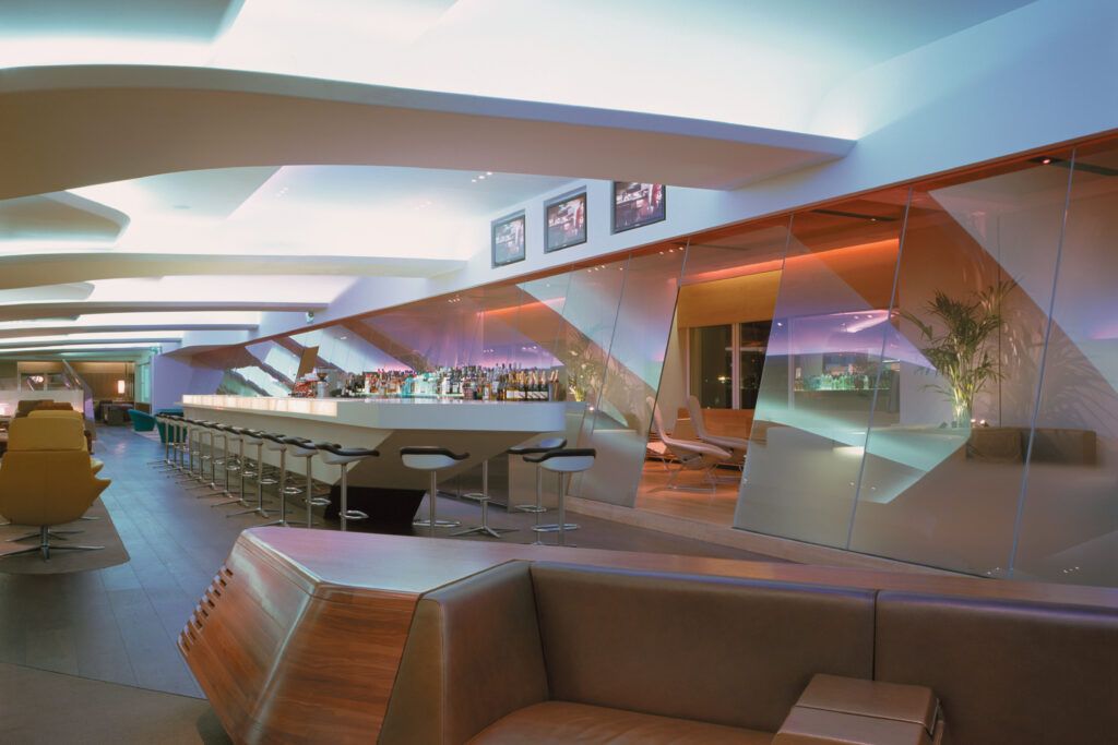 Interior views of the Virgin Atlantic Heathrow Clubhouse in London, designed with a fluid, futuristic aesthetic. The space features organic curves, sculptural ceilings with ambient lighting, and luxurious materials such as wood, leather, and glass. Various zones include a sunken lounge, cocktail bar, restaurant seating with olive trees, and relaxation areas with Eames chairs and large projection screens.