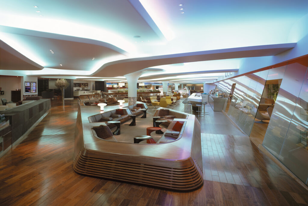 Interior views of the Virgin Atlantic Heathrow Clubhouse in London, designed with a fluid, futuristic aesthetic. The space features organic curves, sculptural ceilings with ambient lighting, and luxurious materials such as wood, leather, and glass. Various zones include a sunken lounge, cocktail bar, restaurant seating with olive trees, and relaxation areas with Eames chairs and large projection screens.