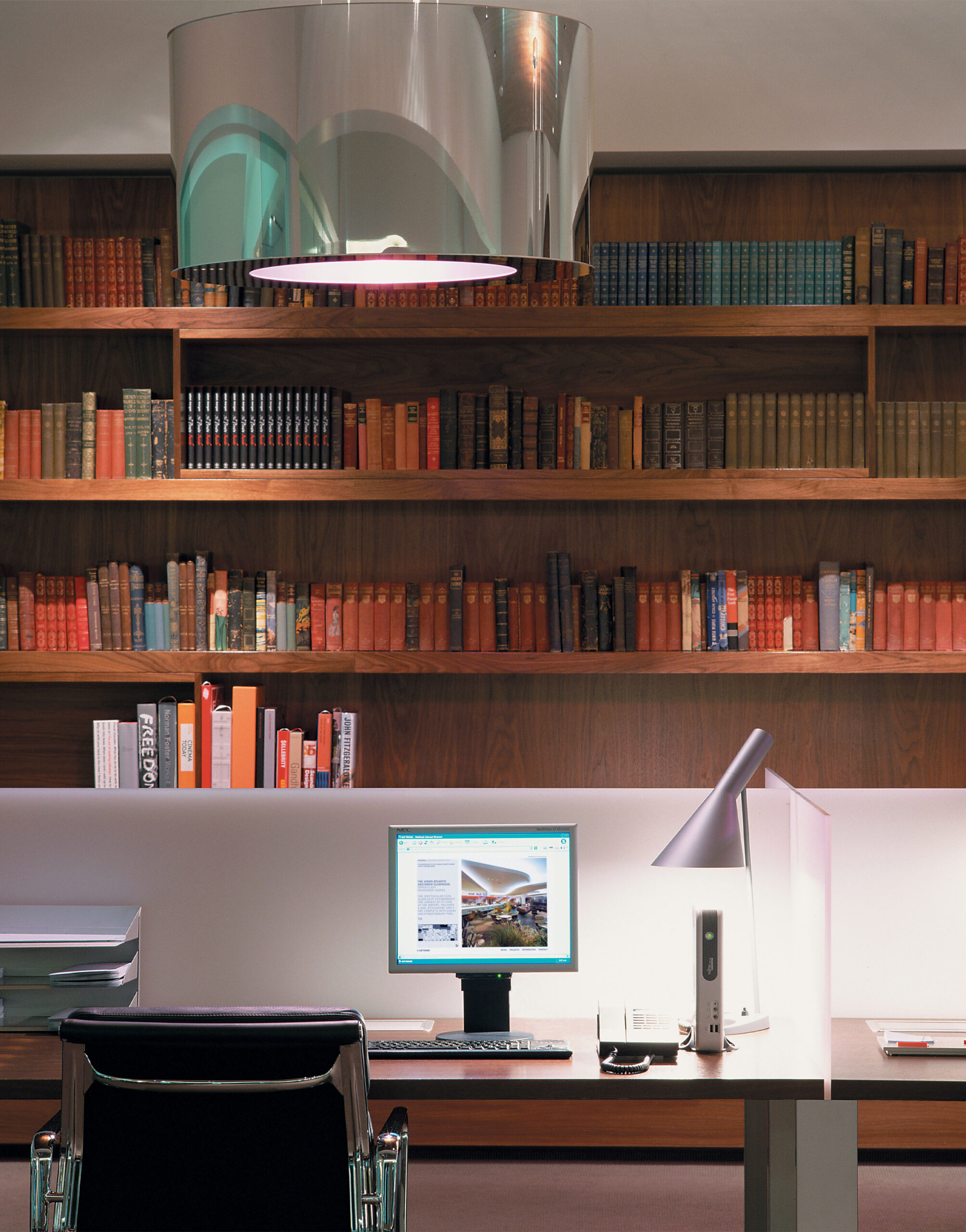 The Office area inside the Virgin Atlantic Heathrow Clubhouse, London — a workspace with a sleek modern desk, computer, and chrome lamp, set against a backdrop of wooden bookshelves filled with colourful hardback books.