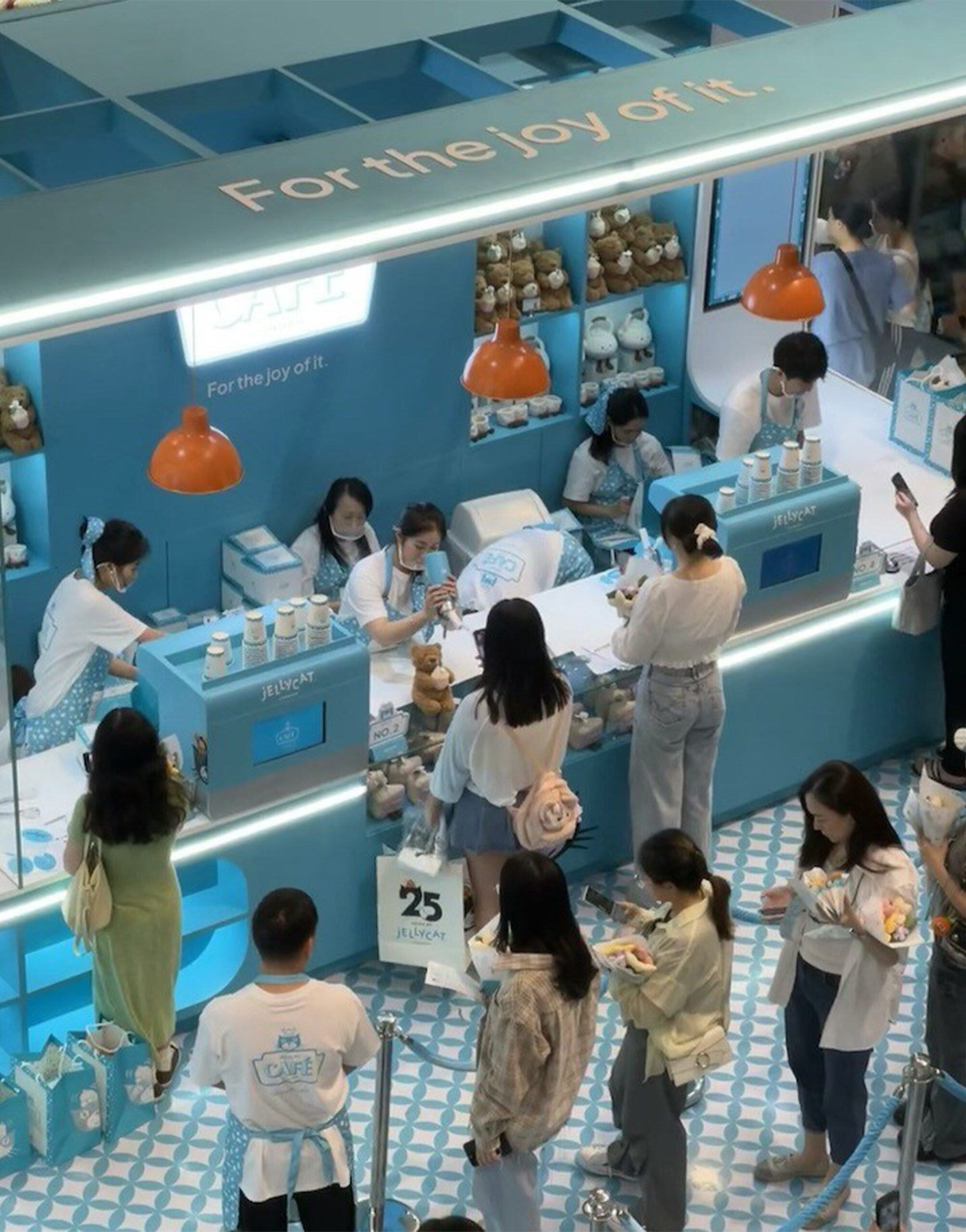 Aerial view of the Jellycat Café pop-up with customers queuing at a bright turquoise counter under signage reading “For the joy of it,” surrounded by shelves of plush toys and playful café décor.