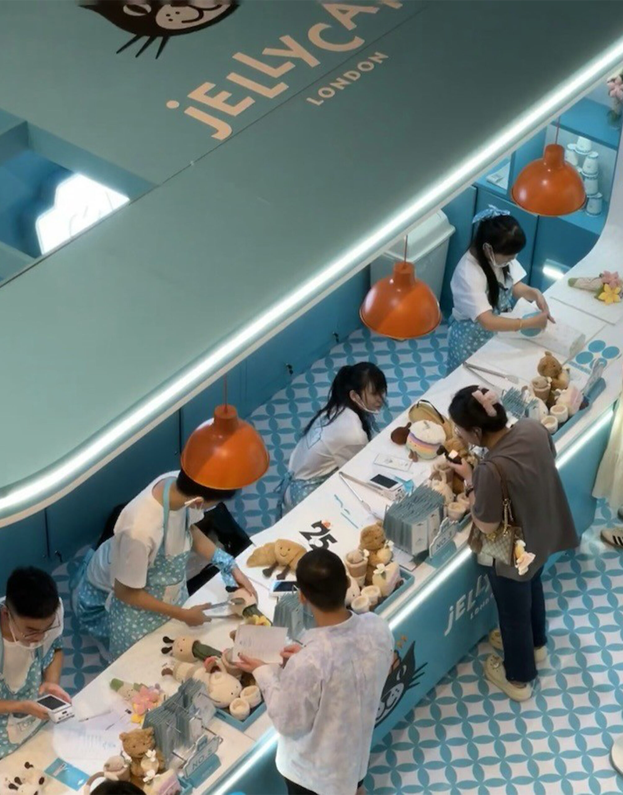 Top-down view of the Jellycat Café pop-up featuring pastel blue interiors, orange pendant lights, and staff assisting customers with plush toy customization on a patterned blue-and-white floor.