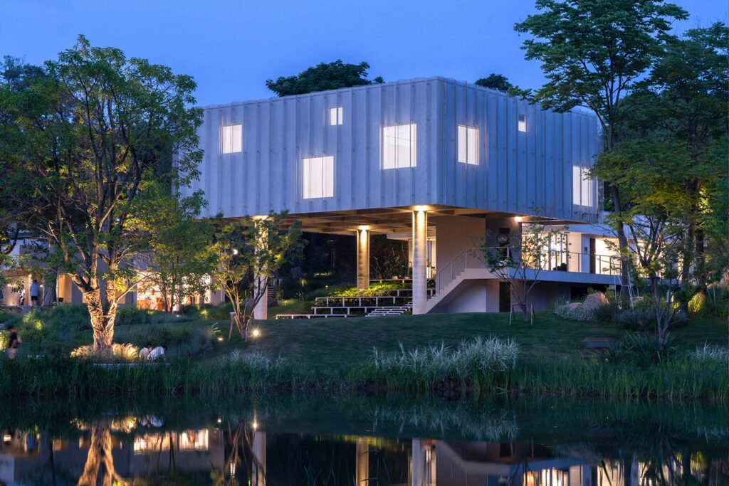 Modern elevated building surrounded by lush greenery and reflected in a serene lake at twilight, featuring minimalist concrete architecture.