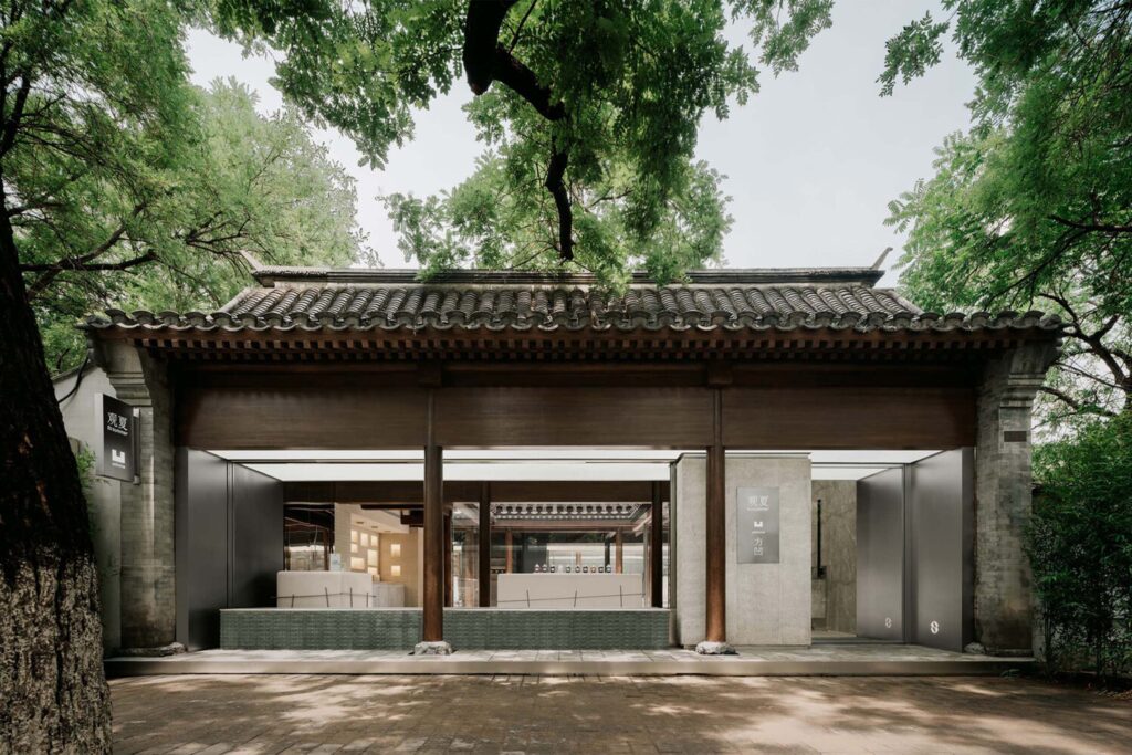Exterior view of To Summer Beijing, blending contemporary design with traditional Chinese courtyard architecture beneath a tiled roof and framed by lush trees.